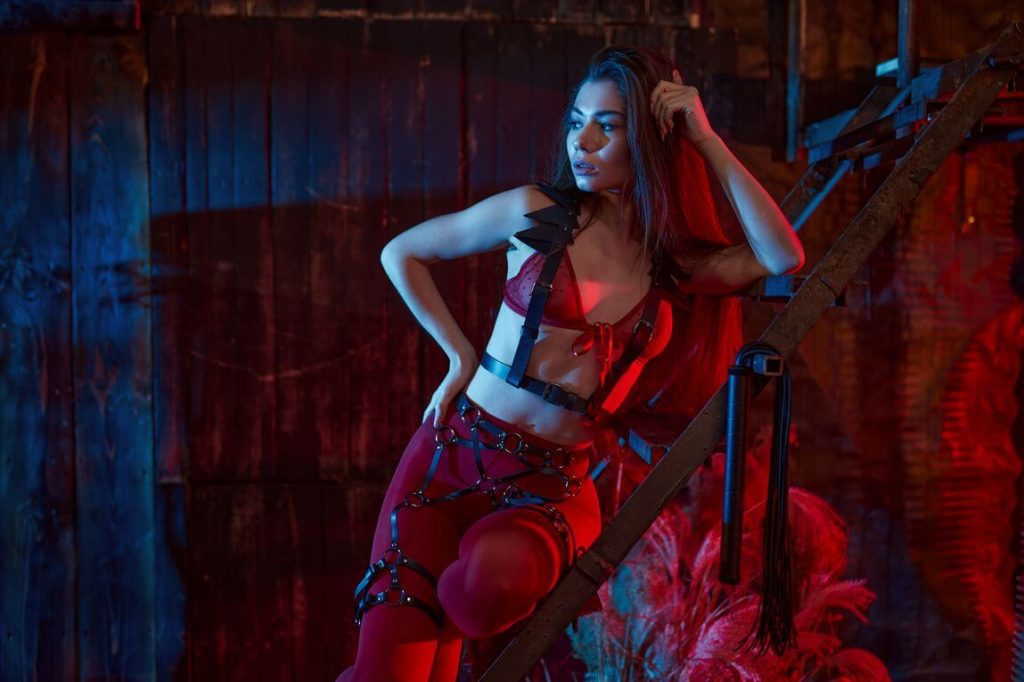 a pornstar wearing a bdsm themed dress while posing inside a room with a red shaded lights