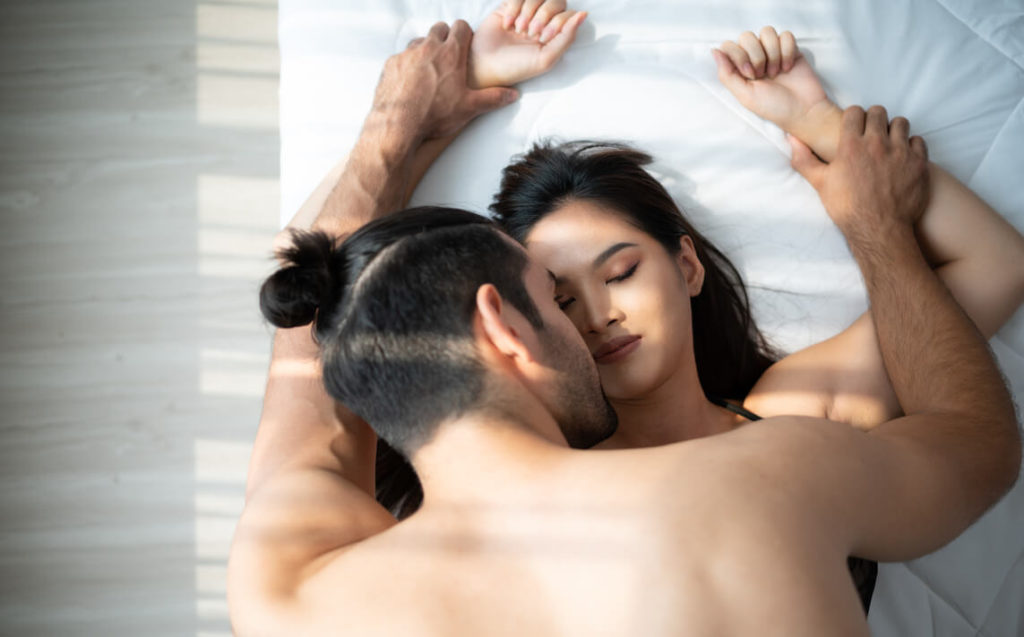 a man with a tied hair holding both of hands of a woman while they are on a bed and having casual encounters